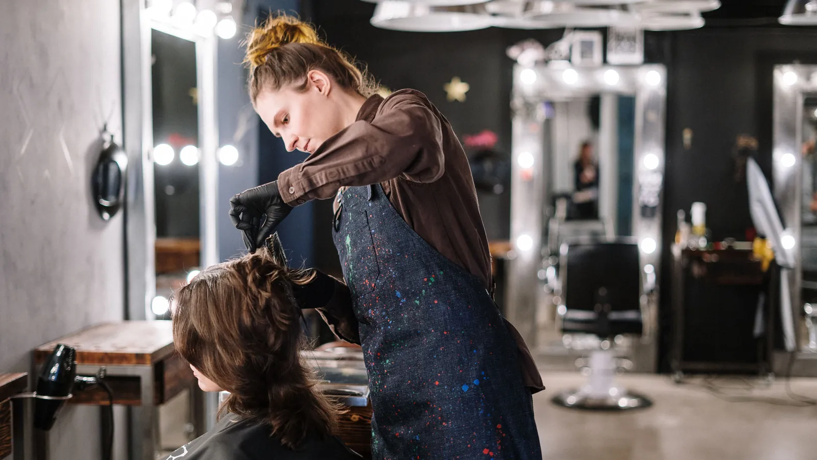 Hairdresser in black gloves working on a client's long hair in a salon with lit mirrors