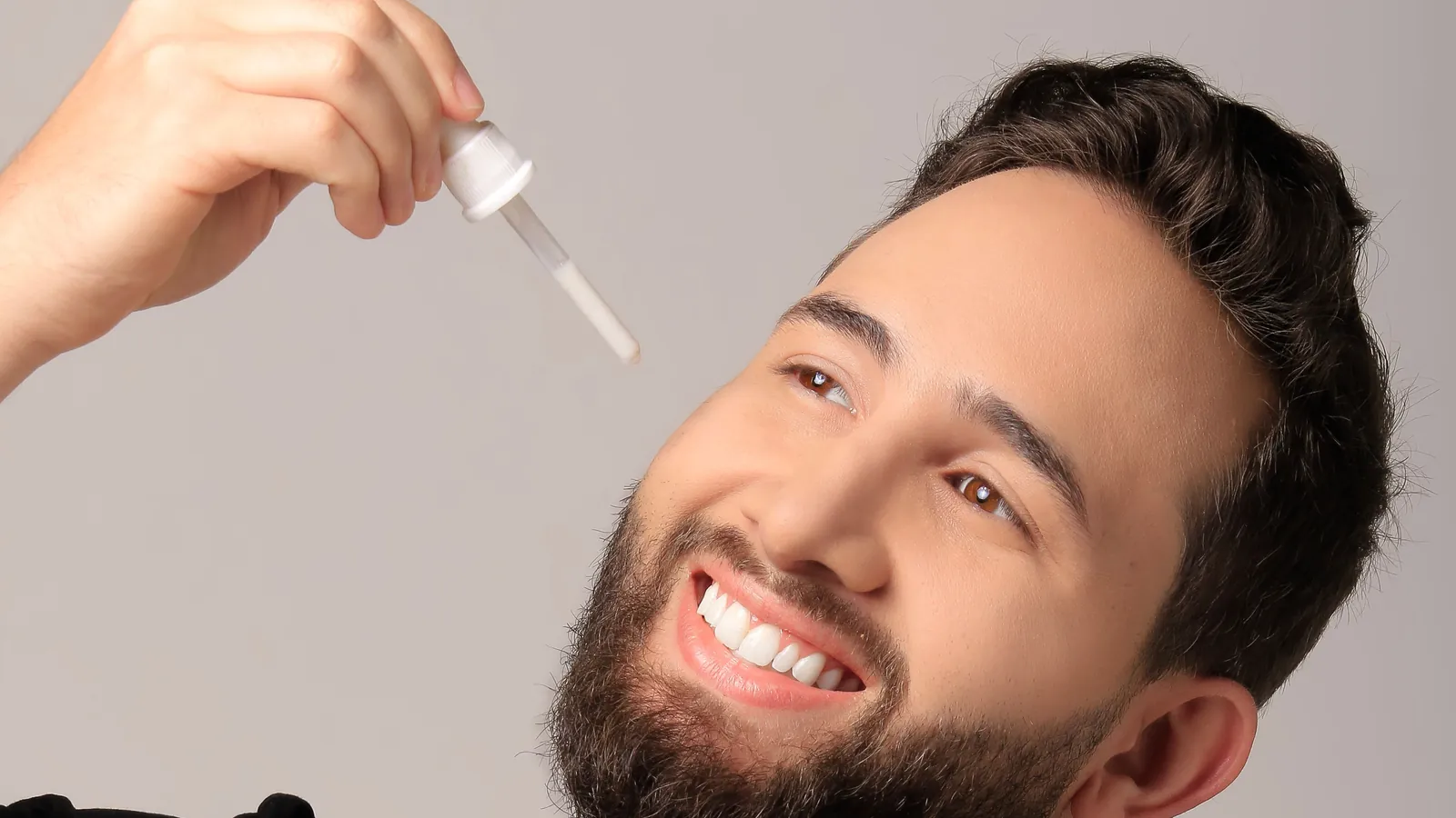 Smiling man with a short beard holding a beard oil dropper