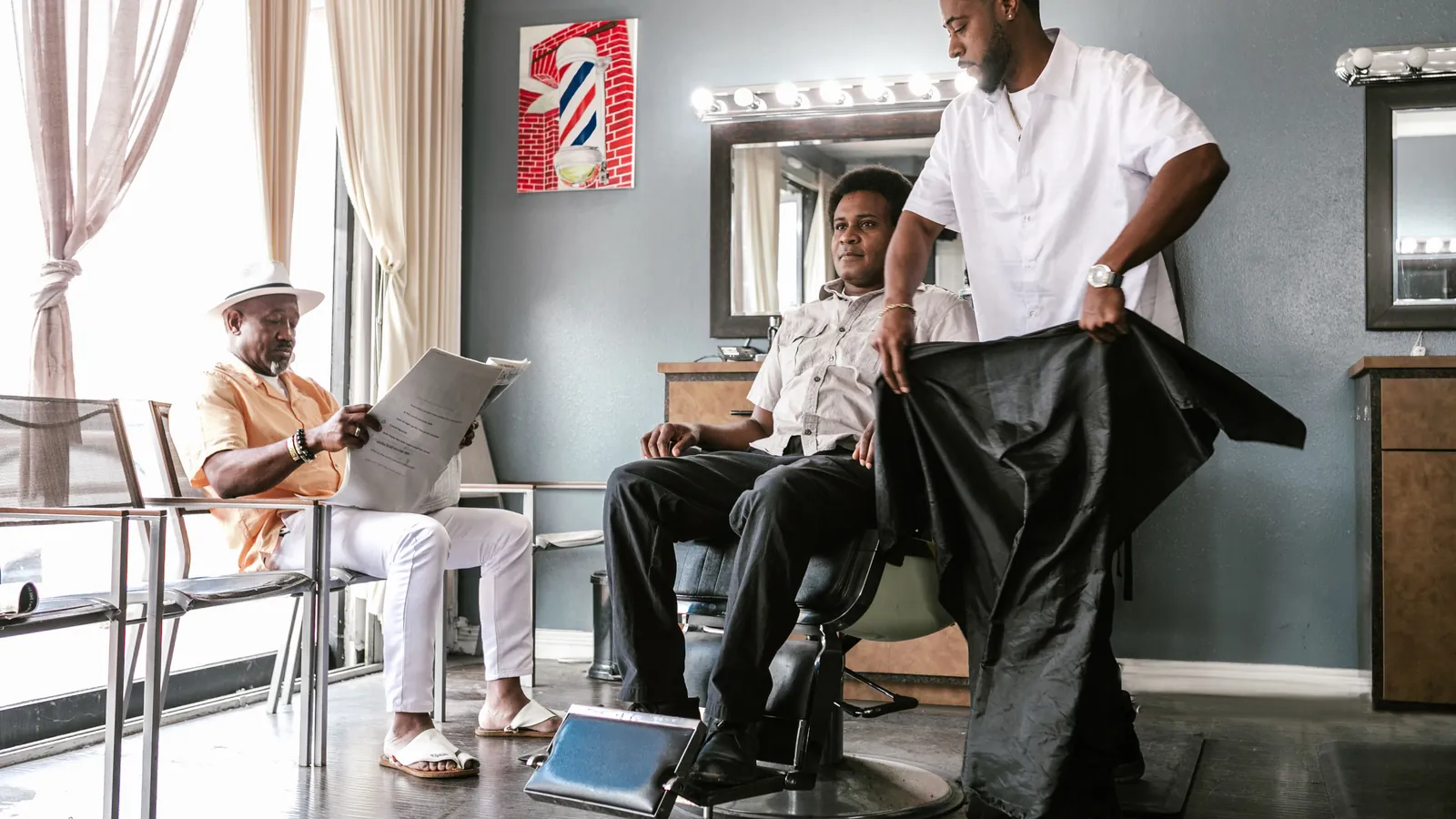 Barber putting a cape on a customer in the chair while another man reads a newspaper in the waiting seats